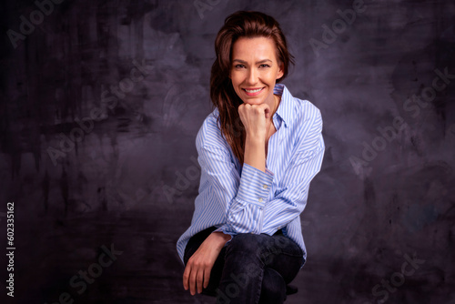 Studio portrait of a brunette haired attractive woman wearing blue shirt and black jeans while posing at isolated dark background