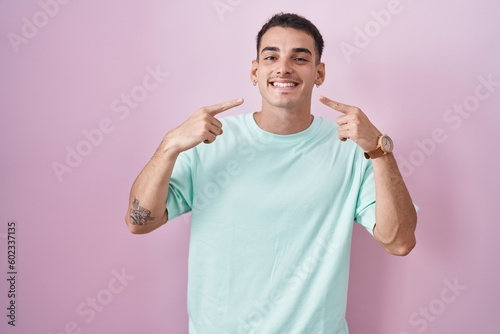 Handsome hispanic man standing over pink background smiling cheerful showing and pointing with fingers teeth and mouth. dental health concept.