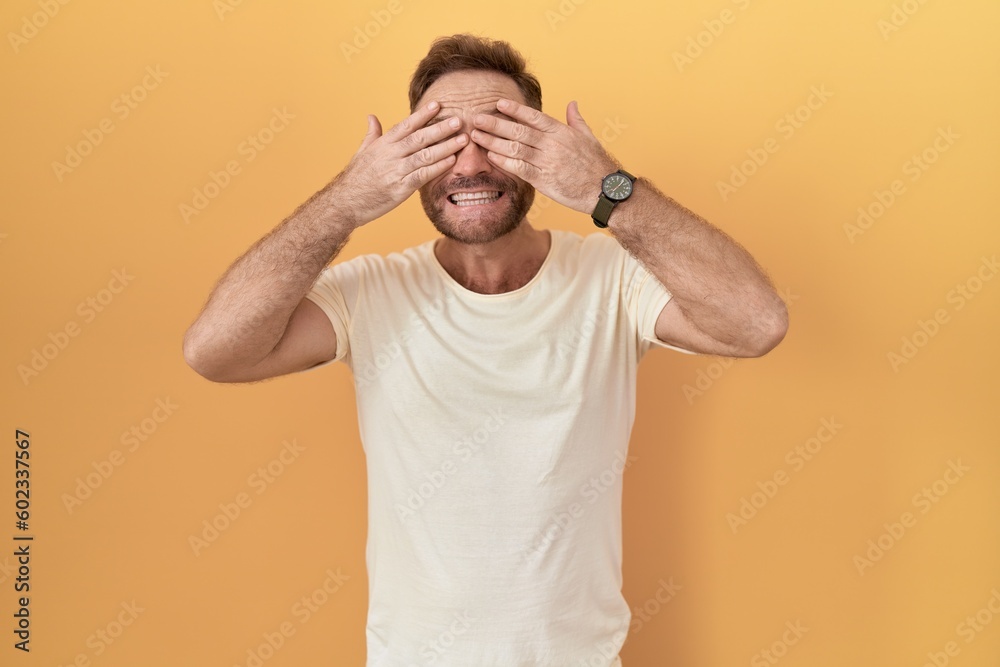 Middle age man with beard standing over yellow background covering eyes with hands smiling cheerful and funny. blind concept. Middle age man with beard standing over yellow background covering eyes with hands smiling cheerful and funny. blind concept.