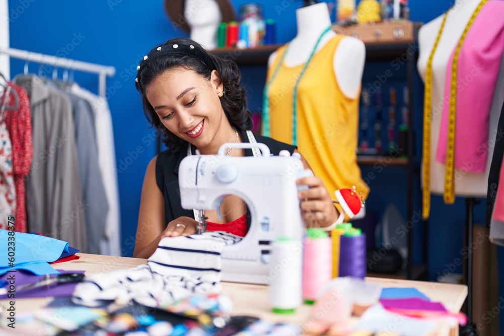 Fototapeta premium Young beautiful hispanic woman tailor smiling confident using sewing machine at clothing factory