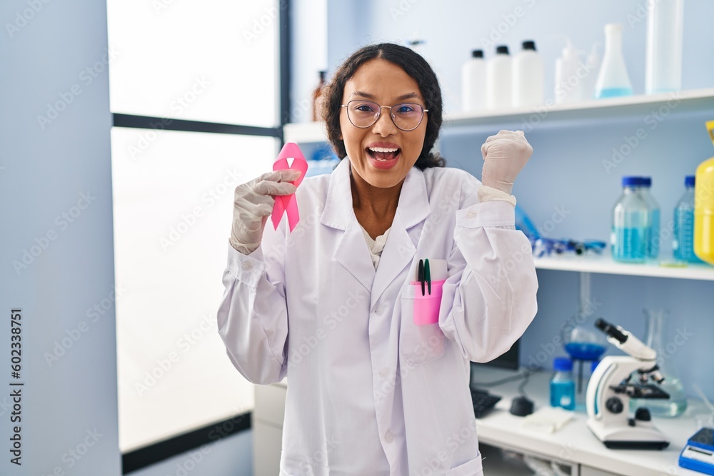 Young hispanic doctor woman working at scientist laboratory holding pink ribbon screaming proud, celebrating victory and success very excited with raised arms Young hispanic doctor woman working at scientist laboratory holding pink ribbon screaming proud, celebrating victory and success very excited with raised arms