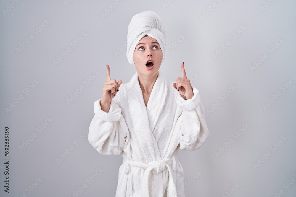 Blonde caucasian woman wearing bathrobe amazed and surprised looking up and pointing with fingers and raised arms. Blonde caucasian woman wearing bathrobe amazed and surprised looking up and pointing with fingers and raised arms.