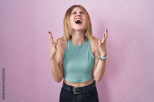 Blonde caucasian woman standing over pink background crazy and mad shouting and yelling with aggressive expression and arms raised. frustration concept.
