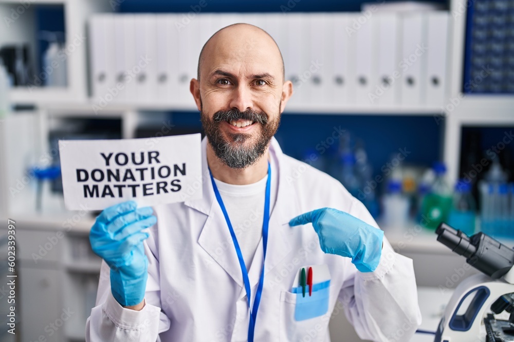 Young hispanic man working at scientist laboratory holding you donation matters banner pointing finger to one self smiling happy and proud