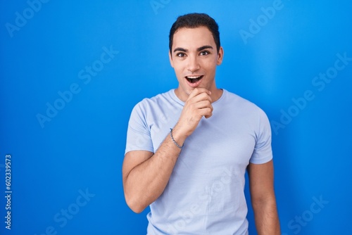 Young hispanic man standing over blue background looking fascinated with disbelief, surprise and amazed expression with hands on chin