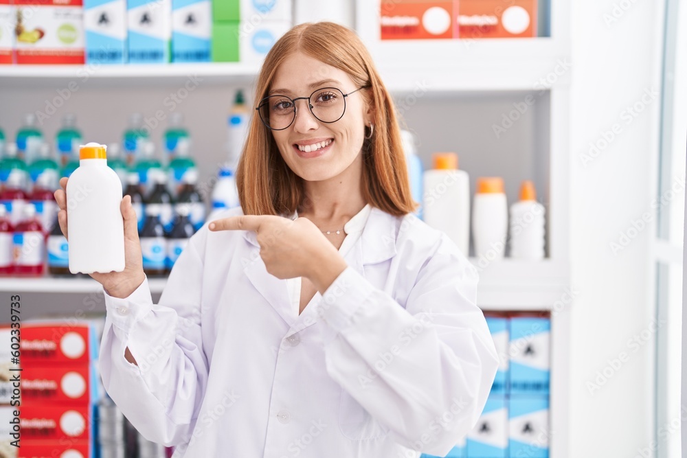 Young redhead woman working at pharmacy drugstore holding sun screen smiling happy pointing with hand and finger