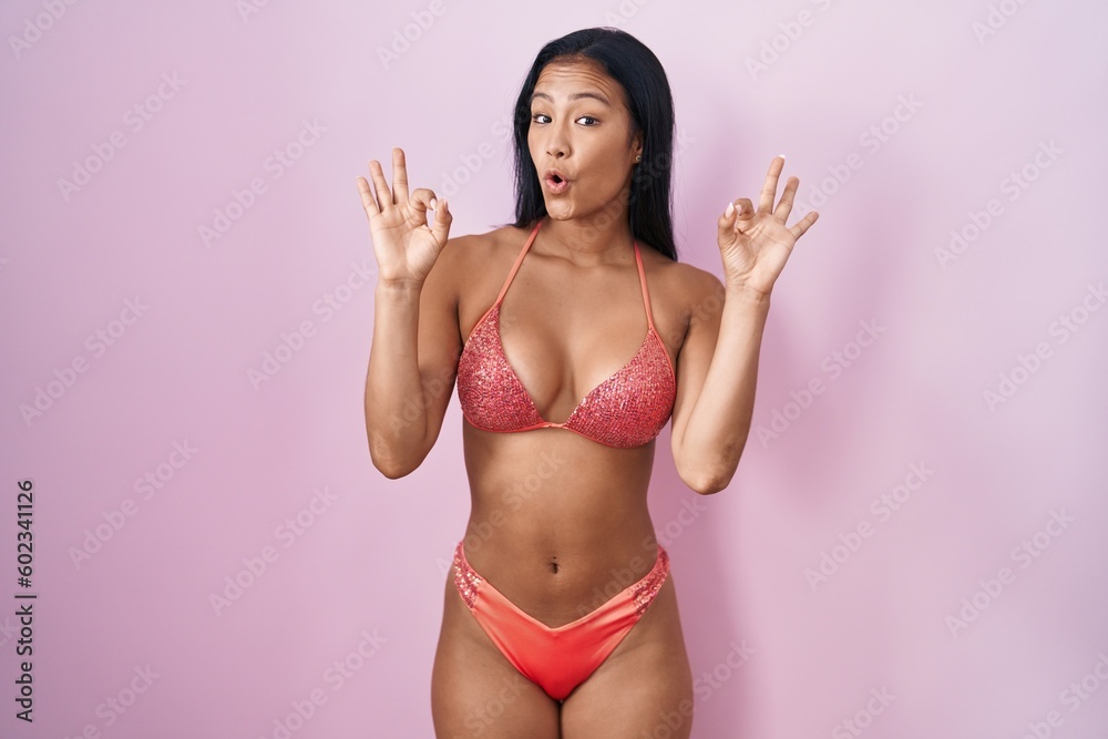 Naklejka premium Hispanic woman wearing bikini looking surprised and shocked doing ok approval symbol with fingers. crazy expression