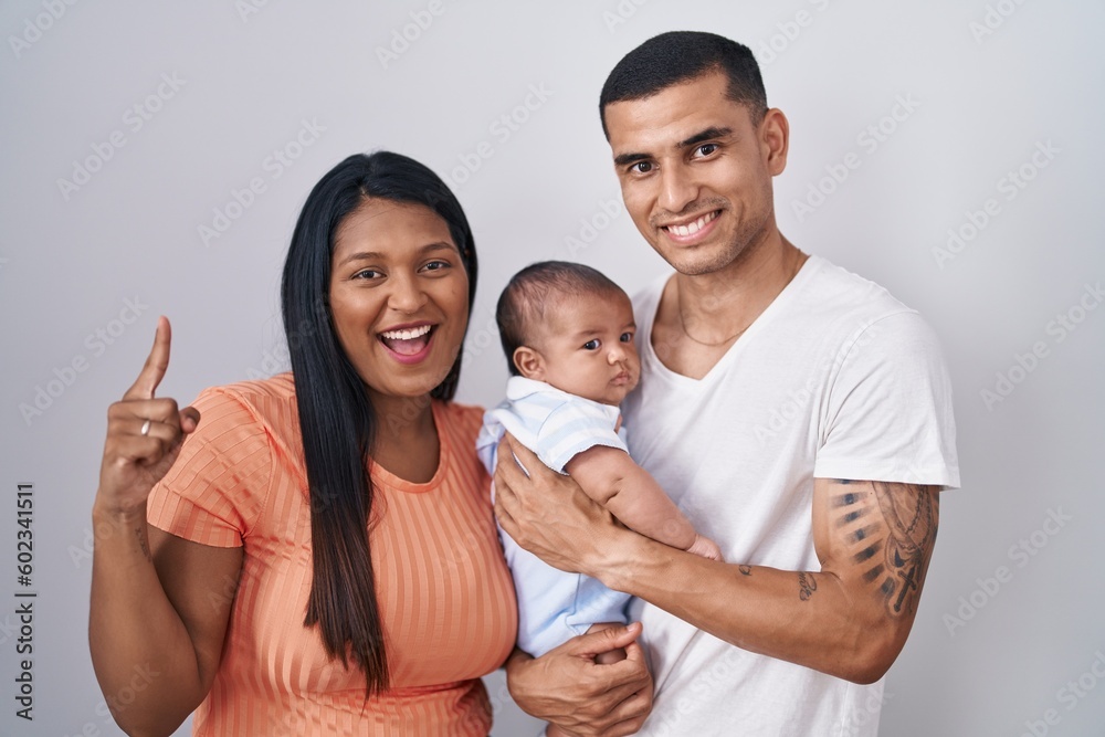 Young hispanic couple with baby standing together over isolated background smiling amazed and surprised and pointing up with fingers and raised arms. Young hispanic couple with baby standing together over isolated background smiling amazed and surprised and pointing up with fingers and raised arms.