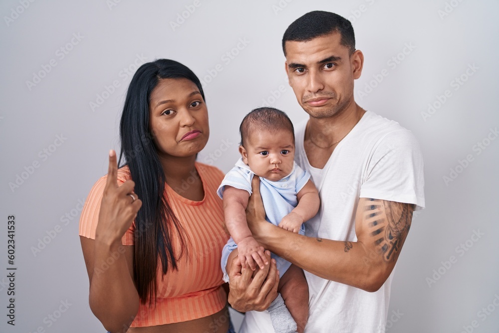 Young hispanic couple with baby standing together over isolated background pointing up looking sad and upset, indicating direction with fingers, unhappy and depressed.