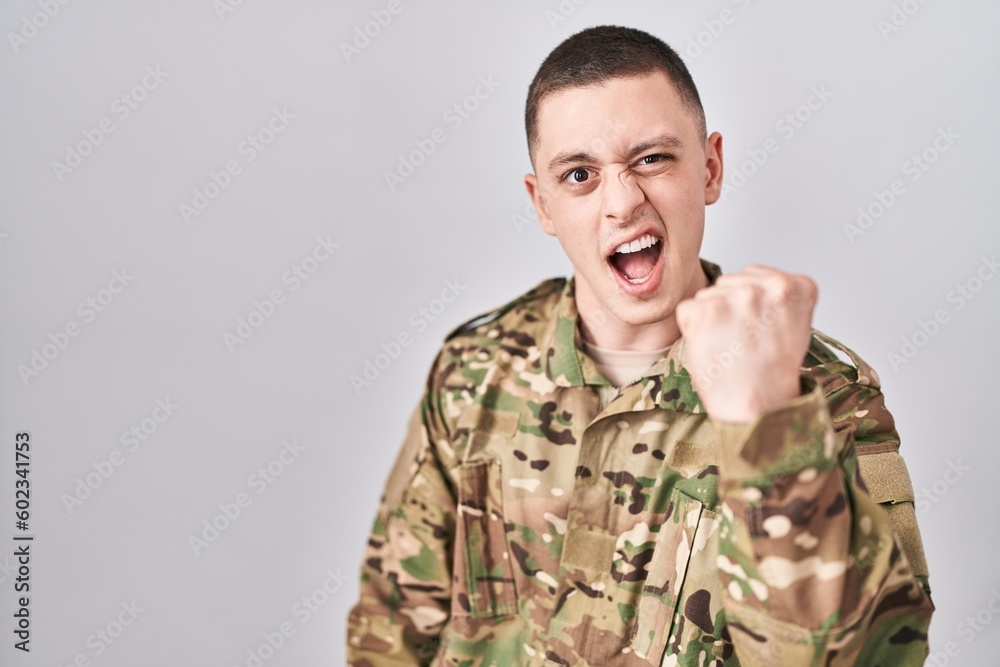 Young man wearing camouflage army uniform angry and mad raising fist frustrated and furious while shouting with anger. rage and aggressive concept.