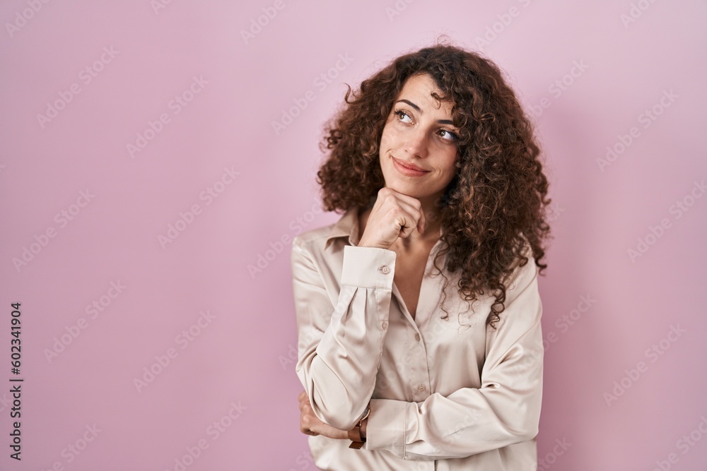 Hispanic woman with curly hair standing over pink background with hand on chin thinking about question, pensive expression. smiling with thoughtful face. doubt concept. Hispanic woman with curly hair standing over pink background with hand on chin thinking about question, pensive expression. smiling with thoughtful face. doubt concept.