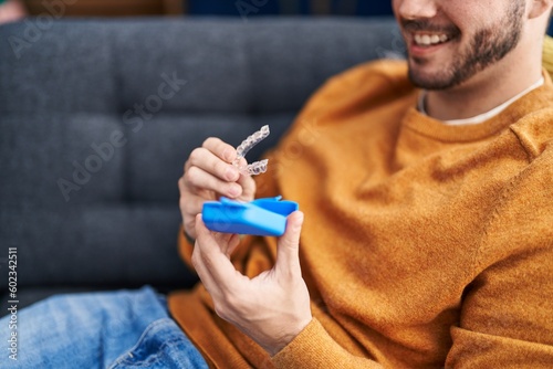 Young hispanic man holding invisible aligner sitting on sofa at home photo