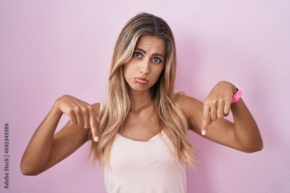 Young blonde woman standing over pink background pointing down looking sad and upset, indicating direction with fingers, unhappy and depressed. Young blonde woman standing over pink background pointing down looking sad and upset, indicating direction with fingers, unhappy and depressed.