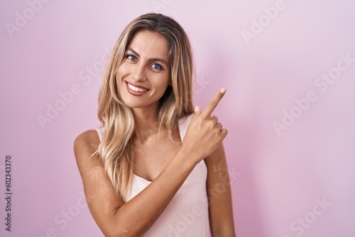 Young blonde woman standing over pink background cheerful with a smile on face pointing with hand and finger up to the side with happy and natural expression