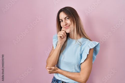 Young hispanic woman standing over pink background looking confident at the camera smiling with crossed arms and hand raised on chin. thinking positive.