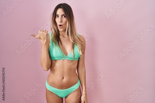 Young hispanic woman wearing bikini over pink background surprised pointing with hand finger to the side, open mouth amazed expression. © Krakenimages.com