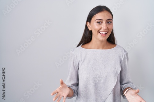Young hispanic woman standing over white background smiling cheerful with open arms as friendly welcome, positive and confident greetings