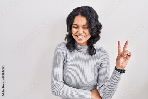 Hispanic woman with dark hair standing over isolated background smiling with happy face winking at the camera doing victory sign with fingers. number two.