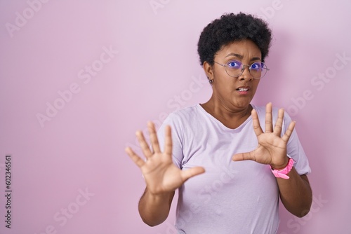 Young african american woman standing over pink background afraid and terrified with fear expression stop gesture with hands, shouting in shock. panic concept.