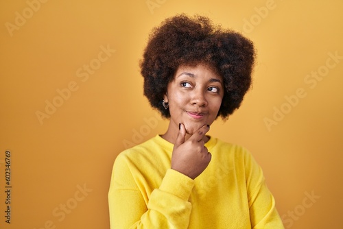 Young african american woman standing over yellow background looking confident at the camera smiling with crossed arms and hand raised on chin. thinking positive.