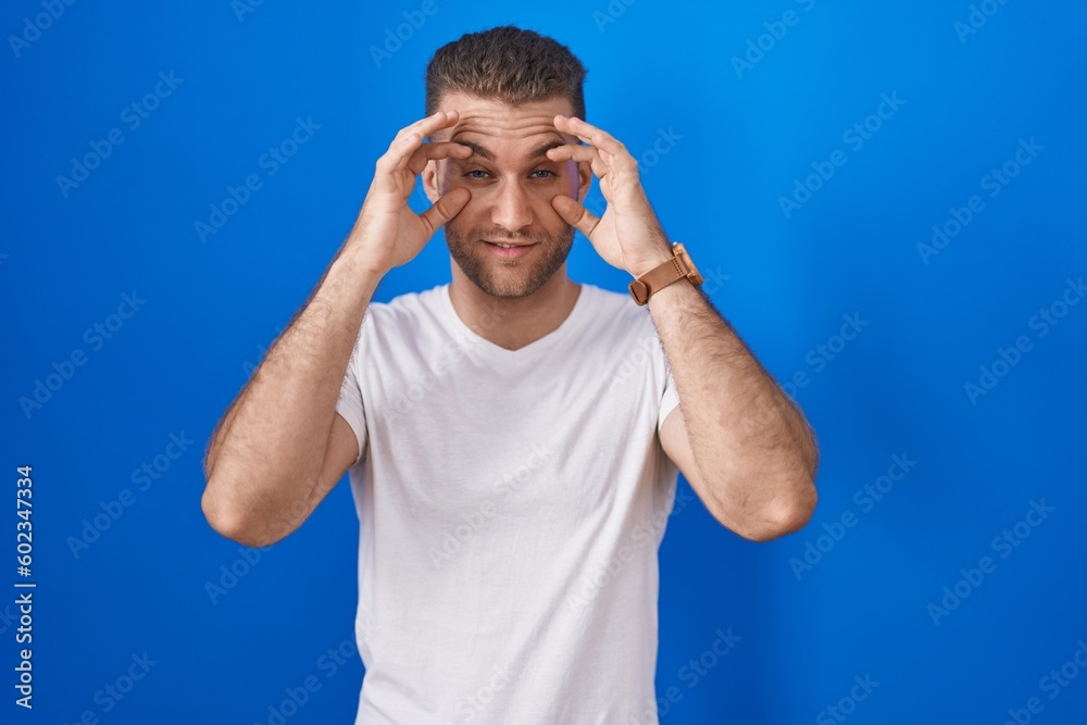 Young caucasian man standing over blue background trying to open eyes with fingers, sleepy and tired for morning fatigue Young caucasian man standing over blue background trying to open eyes with fingers, sleepy and tired for morning fatigue