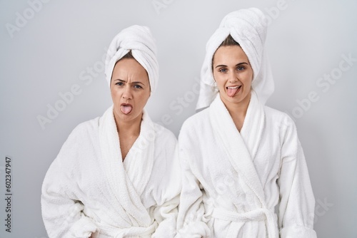Middle age woman and daughter wearing white bathrobe and towel sticking tongue out happy with funny expression. emotion concept.