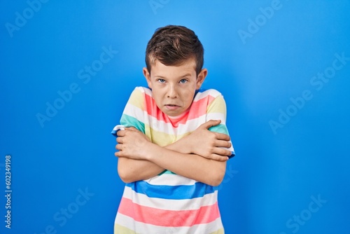 Young caucasian kid standing over blue background shaking and freezing for winter cold with sad and shock expression on face