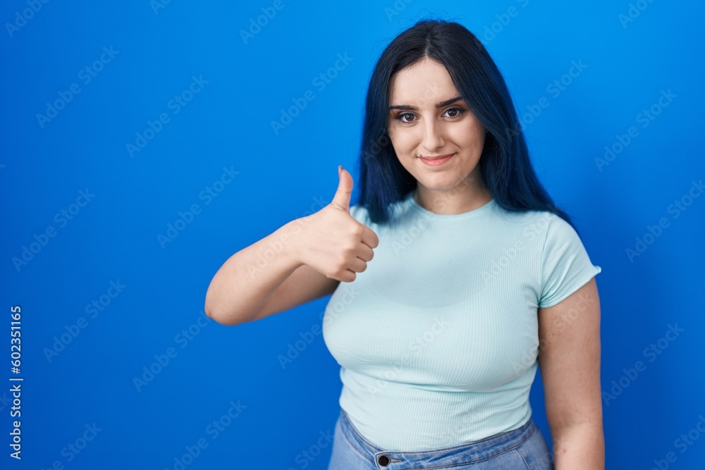 Young modern girl with blue hair standing over blue background doing happy thumbs up gesture with hand. approving expression looking at the camera showing success. Young modern girl with blue hair standing over blue background doing happy thumbs up gesture with hand. approving expression looking at the camera showing success.