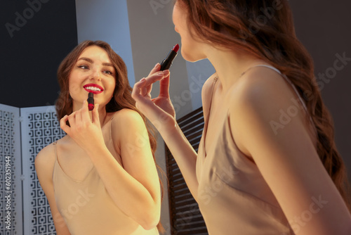 Beautiful young woman in elegant dress applying lipstick near mirror indoors photo