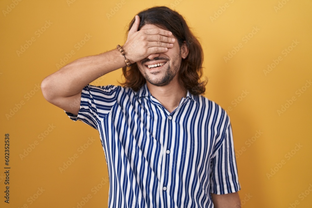 Hispanic man with long hair standing over yellow background smiling and laughing with hand on face covering eyes for surprise. blind concept. Hispanic man with long hair standing over yellow background smiling and laughing with hand on face covering eyes for surprise. blind concept.