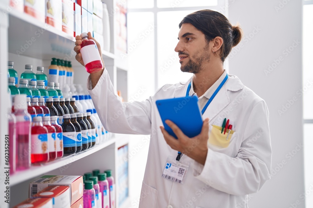 Young hispanic man pharmacist using touchpad holding medication bottle at pharmacy