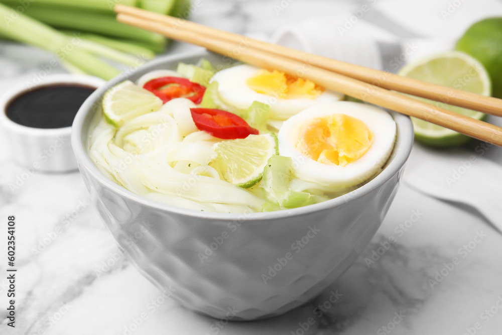 Bowl of delicious rice noodle soup with celery and egg on white marble table, closeup