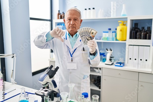 Senior scientist with grey hair working at laboratory holding dollars with angry face, negative sign showing dislike with thumbs down, rejection concept