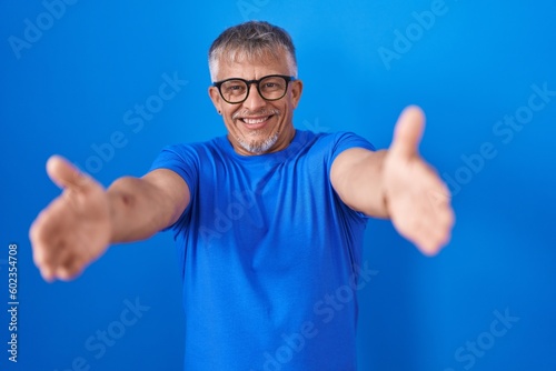 Hispanic man with grey hair standing over blue background looking at the camera smiling with open arms for hug. cheerful expression embracing happiness.
