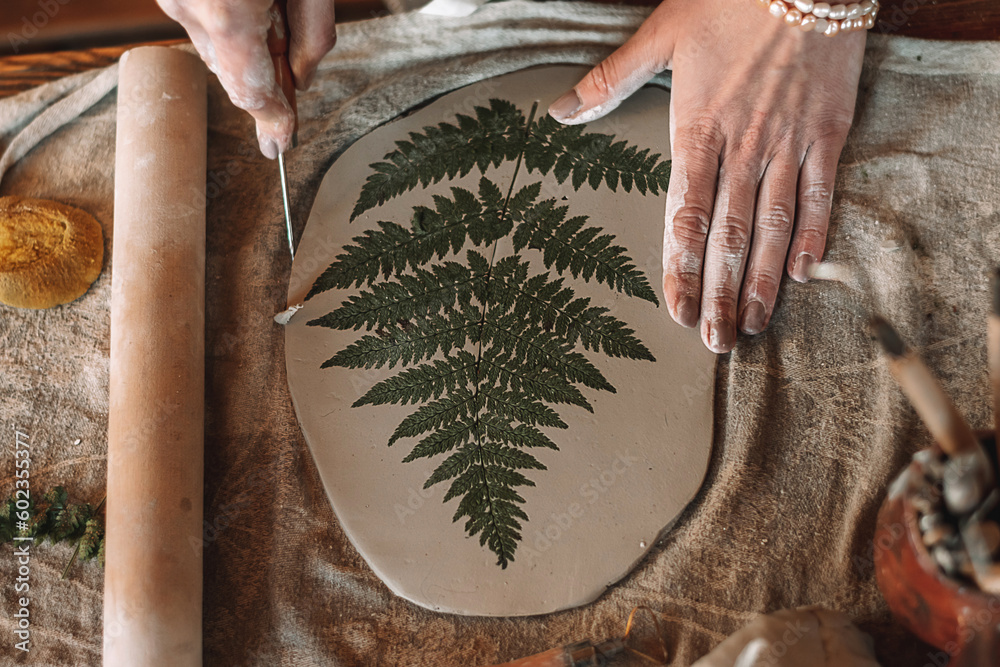 Female sculptor making clay pottery in a home workshop.Making an imprint of a fern leaf on a clay plate,hands close-up.Small business,entrepreneurship,hobby, leisure,sustainability concept.