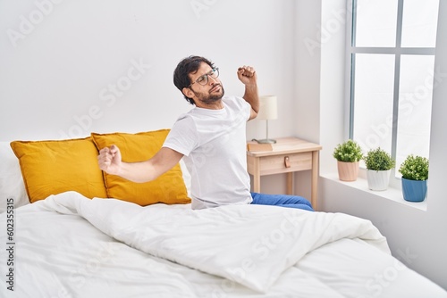 Young hispanic man waking up sitting on bed at bedroom