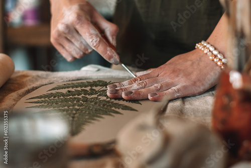 Female sculptor making clay pottery in a home workshop.Making an imprint of a fern leaf on a clay plate,hands close-up.Small business,entrepreneurship,hobby, leisure,sustainability concept.