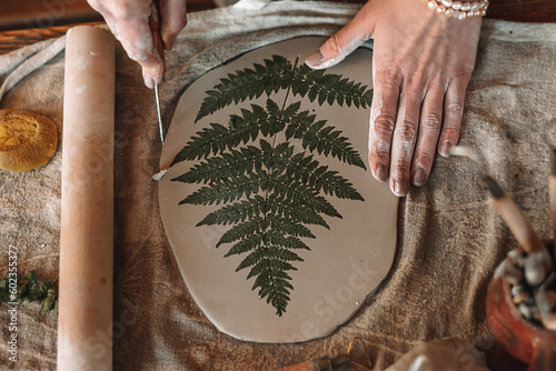 Obraz na płótnie Female sculptor making clay pottery in a home workshop.Making an imprint of a fern leaf on a clay plate,hands close-up.Small business,entrepreneurship,hobby, leisure,sustainability concept. w salonie