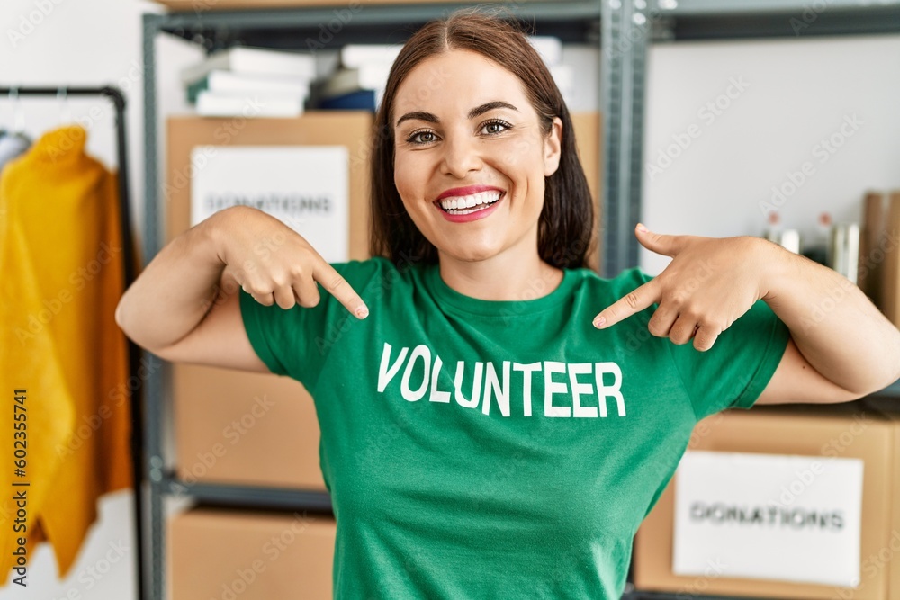 Obraz premium Young beautiful hispanic woman smiling confident pointing with fingers to volunteer t shirt at charity center