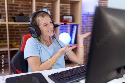 Young caucasian woman playing video games wearing headphones inviting to enter smiling natural with open hand