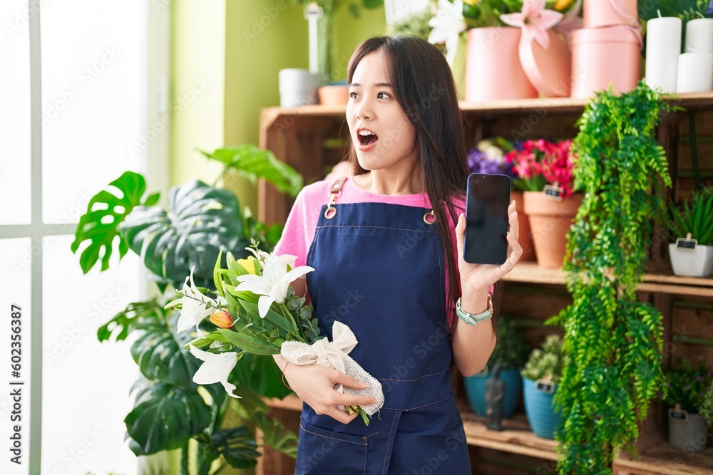 Young chinese woman working at florist shop showing smartphone screen angry and mad screaming frustrated and furious, shouting with anger. rage and aggressive concept.