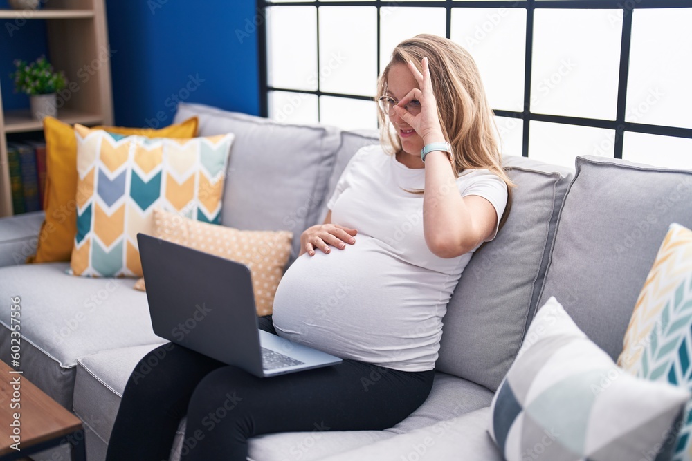 Young pregnant woman sitting on the sofa at home using laptop smiling happy doing ok sign with hand on eye looking through fingers Young pregnant woman sitting on the sofa at home using laptop smiling happy doing ok sign with hand on eye looking through fingers