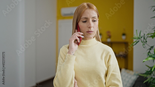Young blonde woman talking on smartphone with serious expression at home