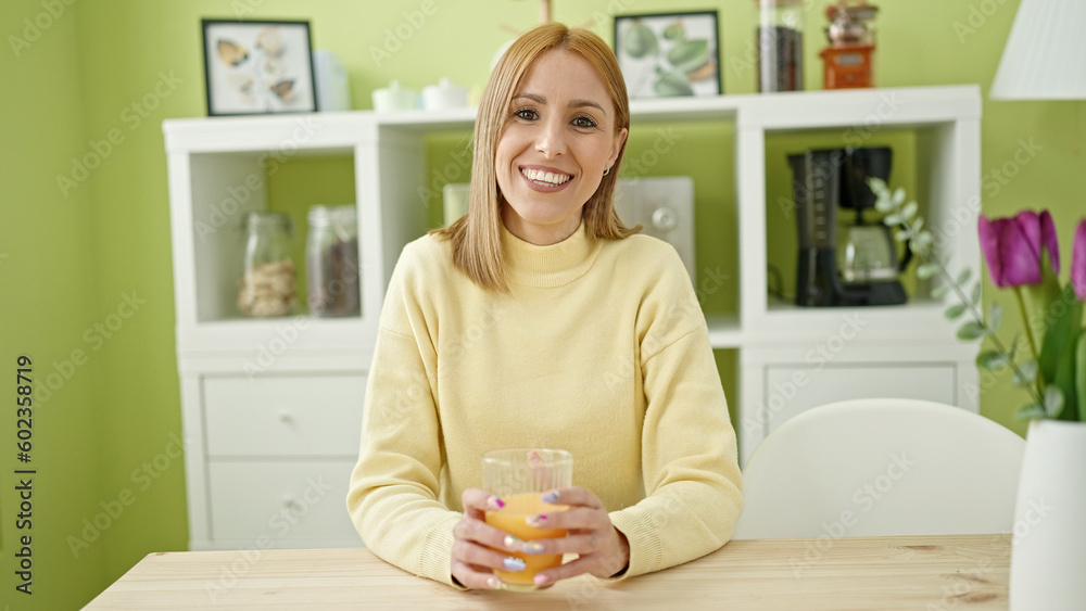Young blonde woman drinking orange juice sitting on table at home