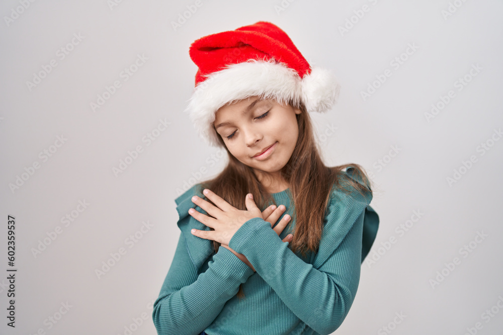 Little caucasian girl wearing christmas hat smiling with hands on chest, eyes closed with grateful gesture on face. health concept. Little caucasian girl wearing christmas hat smiling with hands on chest, eyes closed with grateful gesture on face. health concept.
