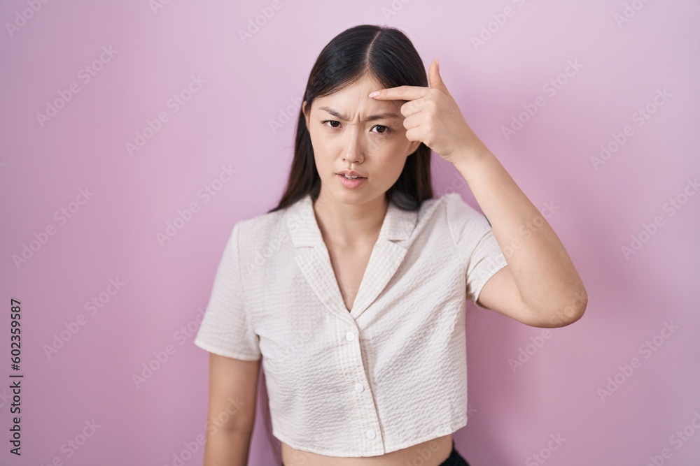 Chinese young woman standing over pink background pointing unhappy to pimple on forehead, ugly infection of blackhead. acne and skin problem Chinese young woman standing over pink background pointing unhappy to pimple on forehead, ugly infection of blackhead. acne and skin problem