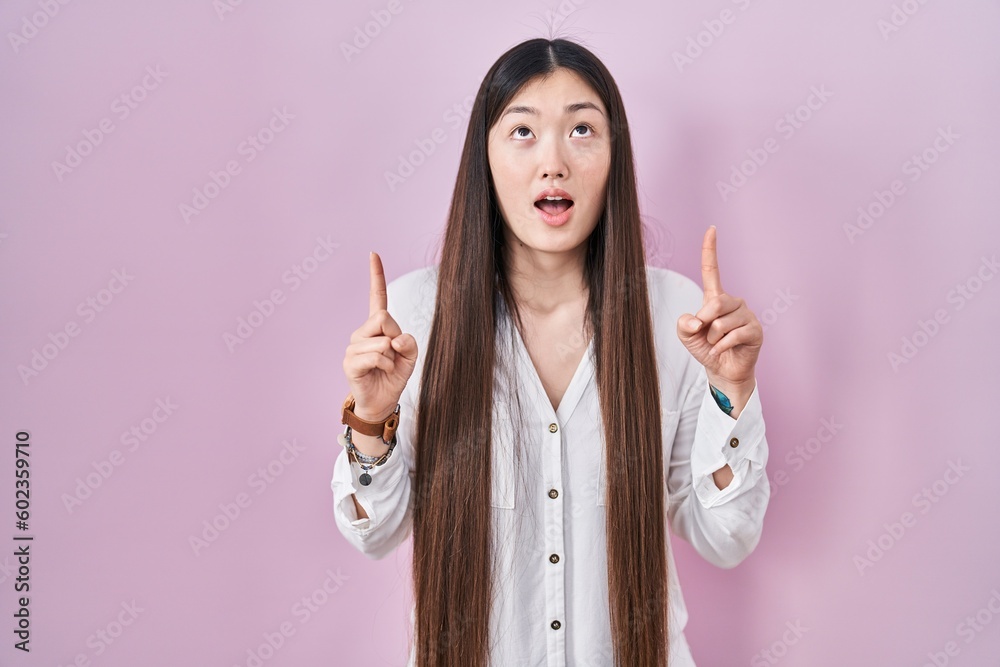 Chinese young woman standing over pink background amazed and surprised looking up and pointing with fingers and raised arms. Chinese young woman standing over pink background amazed and surprised looking up and pointing with fingers and raised arms.