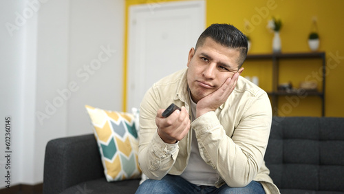 Young hispanic man watching tv sitting on sofa at home