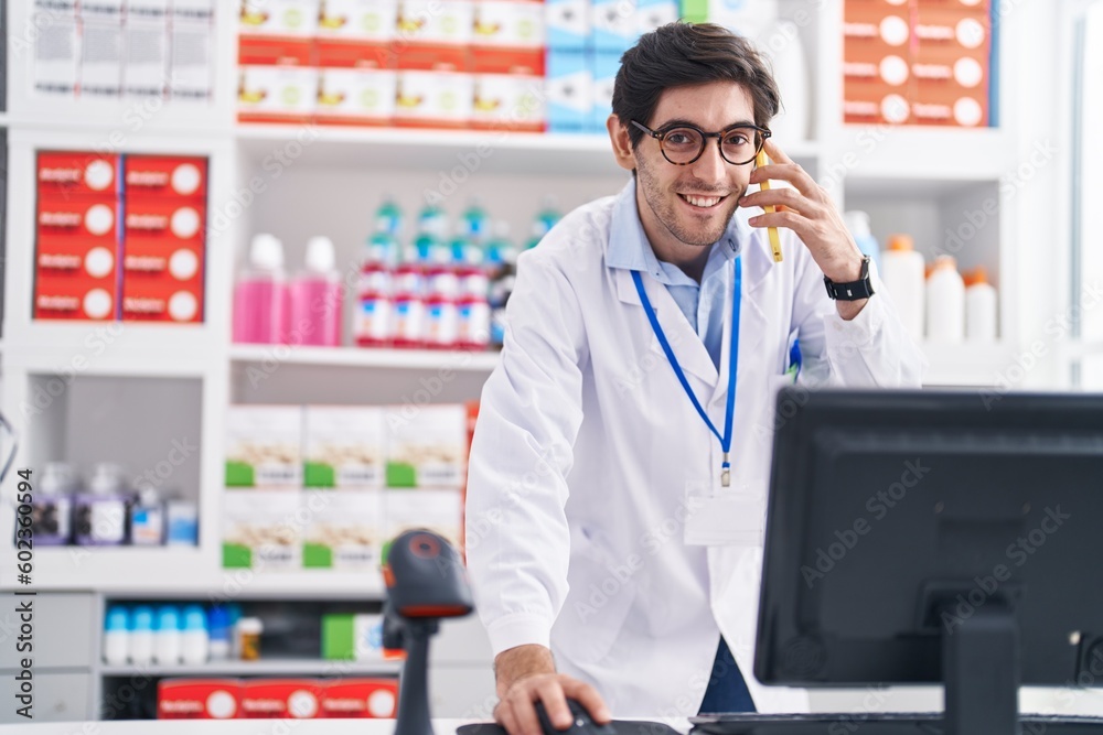 Young hispanic man pharmacist talking on smartphone using computer at pharmacy