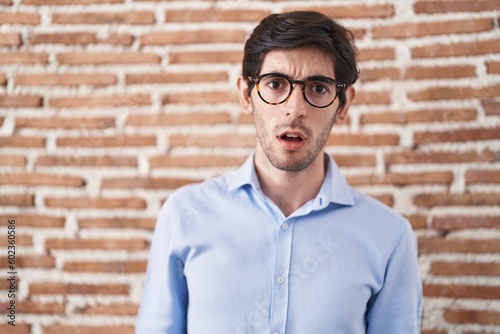 Young hispanic man standing over brick wall background in shock face, looking skeptical and sarcastic, surprised with open mouth